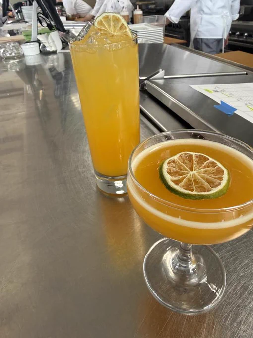 Two cocktails on a metal counter. One is a tall glass with an orange drink and a dried lemon slice. The other is a coupe glass with an orange cocktail, garnished with a slice of lime and lemon. A kitchen setup is visible in the background.