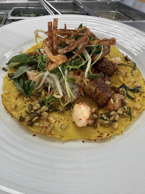 A vibrant dish featuring a golden Vietnamese bánh xèo pancake topped with shrimp, pork, and an array of fresh greens and sprouts. Crispy fried strips are garnished on top, adding texture. The dish is served on a white plate.