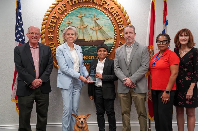 A group of six people is standing in front of the City of Tampa, Florida seal. They are posing for a photo with one person holding a small trophy. A small dog is sitting in the center. The American flag and another flag are visible in the background.