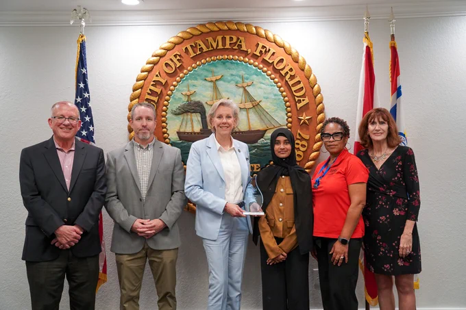 A group of six people is standing in front of the City of Tampa, Florida seal. They are posing for a photo with one person holding a small trophy. A small dog is sitting in the center. The American flag and another flag are visible in the background.