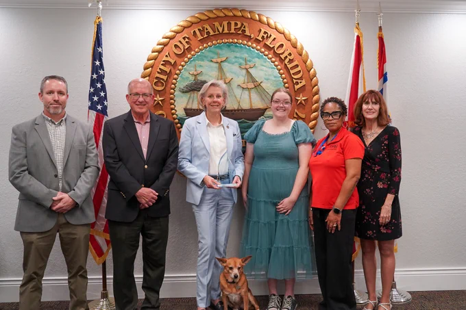 A group of six people is standing in front of the City of Tampa, Florida seal. They are posing for a photo with one person holding a small trophy. A small dog is sitting in the center. The American flag and another flag are visible in the background.