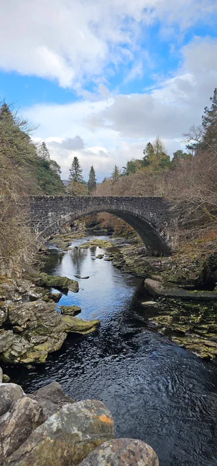 A stone bridge above a small stream with a patch of blue sky reflecting in the water.