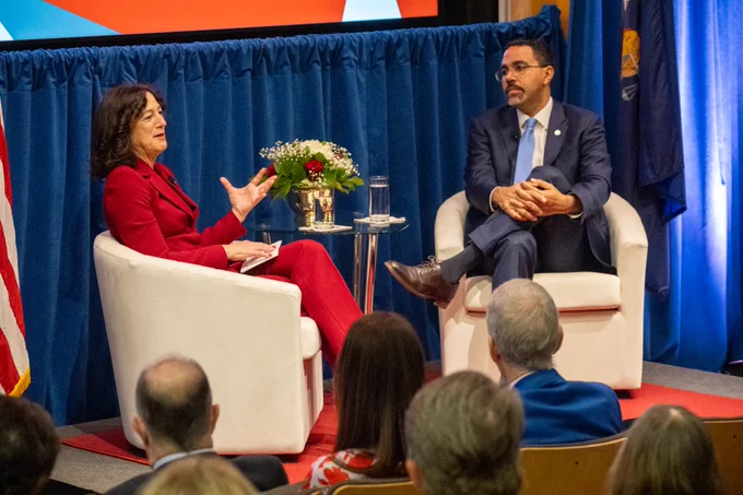 Stony Brook Univeristy President Andrea Goldsmith weas a red suit and SUNY Chancelor John B. King Jr. wears a dark blue suit are seated in white chairs, engaged in conversation on a stage. There is a small table with a floral arrangement between them. A U.S. flag and a blue curtain are in the background. An audience is visible in the foreground.