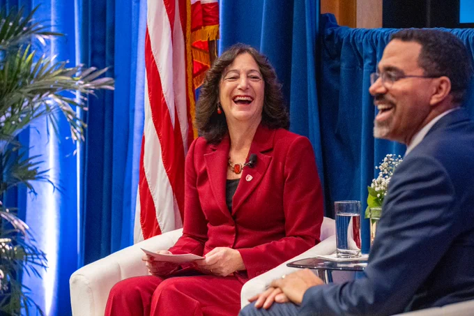 Stony Brook Univeristy President Andrea Goldsmith weas a red suit and SUNY Chancelor John B. King Jr. wears a dark blue suit are seated in white chairs. They are smiling and engaged in conversation. An American flag is visible in the background. A potted plant and a glass of water are by their side.