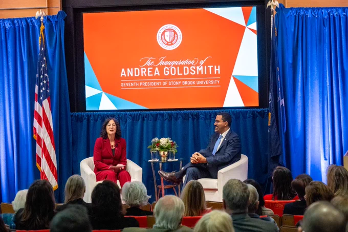 Stony Brook Univeristy President Andrea Goldsmith weas a red suit and SUNY Chancelor John B. King Jr. wears a dark blue suit are seated in white chairs on a stage with a backdrop featuring "The Inauguration of Andrea Goldsmith, Seventh President of Stony Brook University." The setting includes an audience, U.S. and New York State flags, and a floral arrangement.