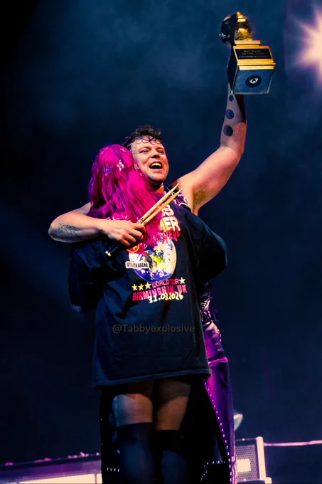 An image of Ashton Irwin hugging a fan with one arm that is holding drum sticks while the other hand holds up a fake boyband of the year trophy 