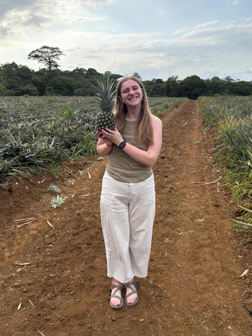 A person stands on a dirt path in a pineapple field, holding a pineapple with both hands. They are smiling, wearing a sleeveless top, light-colored pants, and sandals. The sky is overcast, and trees are visible in the background.