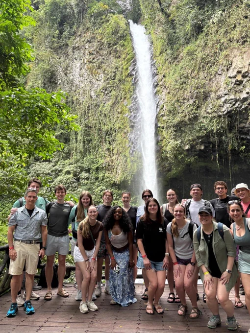 A group of people standing in front of a tall, flowing waterfall surrounded by lush greenery.
