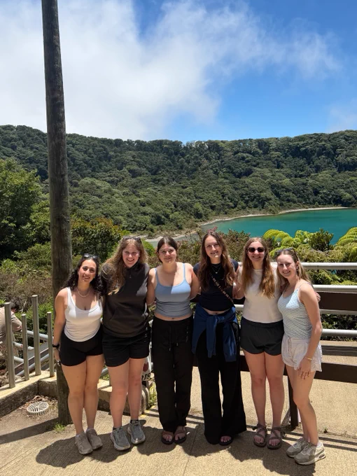 A group of six smiling people standing together on a pathway with lush greenery and a turquoise lake in the background. They are dressed casually and appear to be enjoying a sunny day outdoors.
