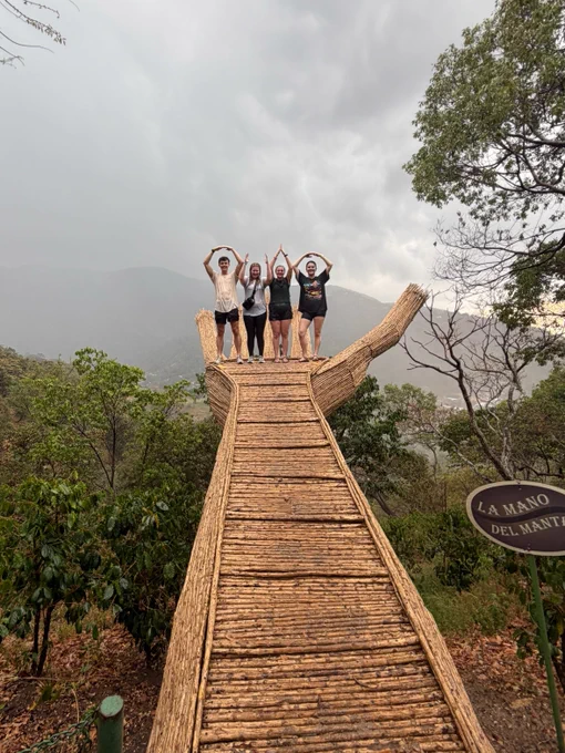Four people stand on a large wooden sculpture resembling a hand, with arms raised in the air. The background shows a misty, mountainous landscape with lush greenery. A sign to the side reads "La Mano del Mártir.