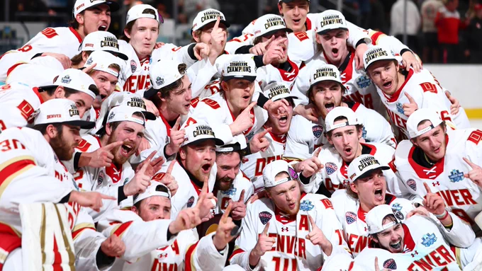 A group of Denver hockey team players wearing white uniforms and hats with "Champions" written on them celebrate on the ice. They are smiling and pointing, with excitement and camaraderie evident.