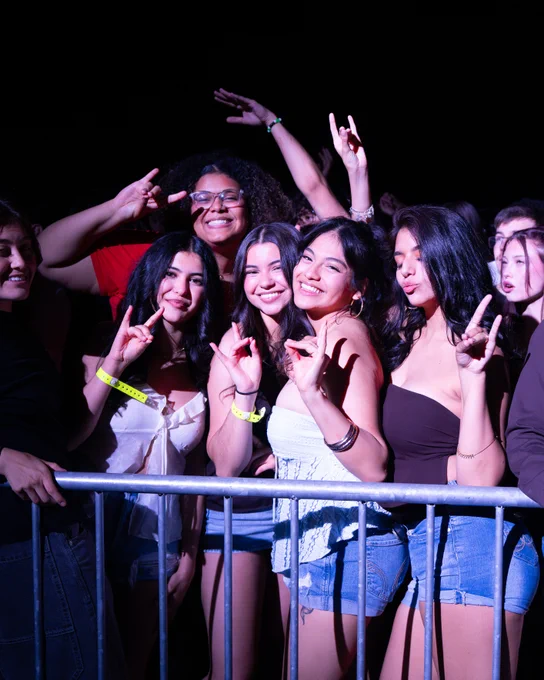 A group of people smiling and posing for a photo at Fiestas, standing in front of a metal barrier. They are making the Lobo hand sign. It is nighttime, and the area is dimly lit, highlighting their faces.