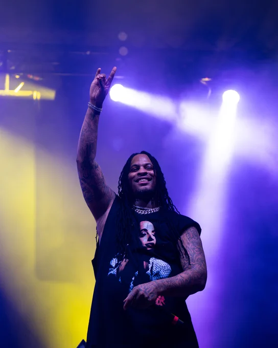 Waka Flocka Flame holds up the Lobo hand sign. The background is illuminated with purple and yellow stage lights.