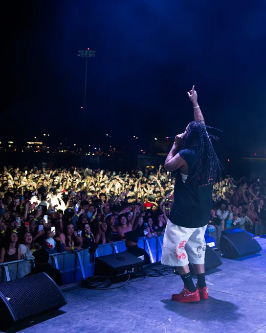 Waka Flocka Flame on stage facing a large, energetic crowd at night, with outstretched arm pointing upward. Audience is gathered under night sky with concert lights in background.