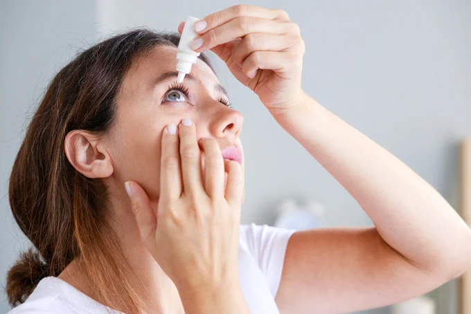 We see the image of a woman preparing to self-administer an eye drop into her right eye.