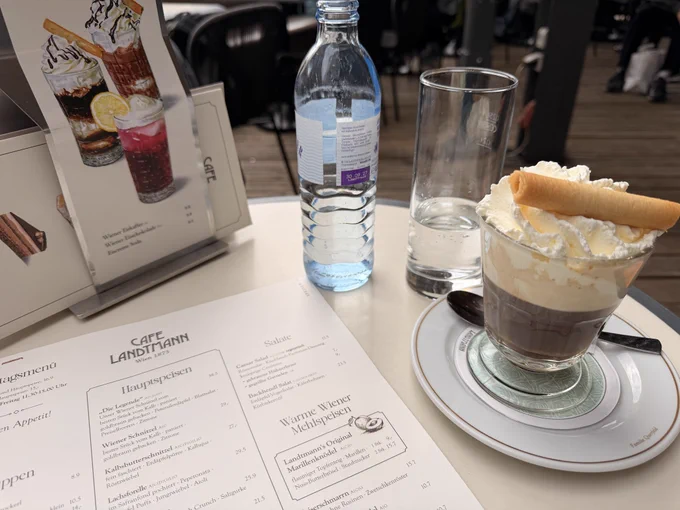 A cup of coffee with whipped cream, a small bottle of water and a drinking glass on a table top