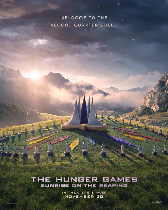 Promotional graphic for The Hunger Games: Sunrise on the Reaping announcing the trailer release. A scenic green valley with mountains in the background features a circular floral installation arranged in colorful sections around a central geometric structure, with people seated evenly around the display.