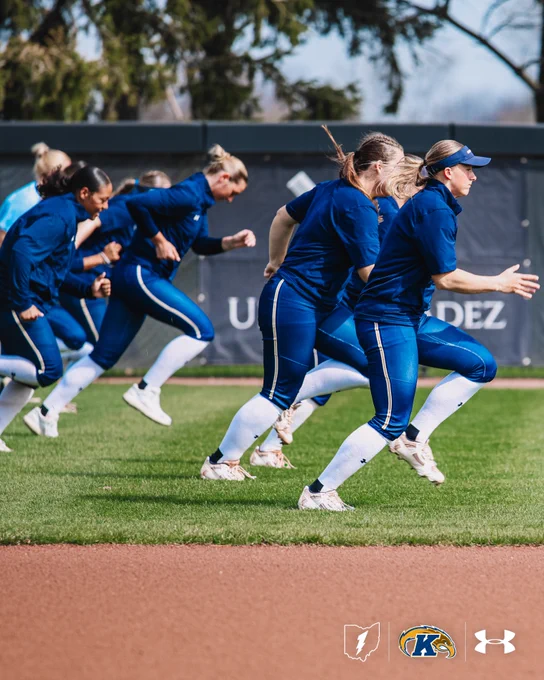 Kent State softball players run sprints along the outfield grass during warm-ups, captured mid-stride in a dynamic action shot. All players wear navy blue uniforms with white knee-high socks.