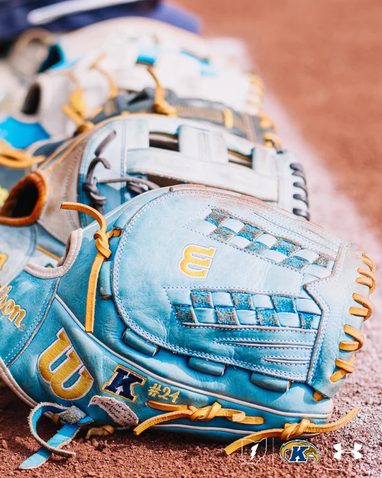 Three light blue Wilson softball gloves with gold lacing and 'KU' branding, including one labeled '#24,' rest on the infield dirt along the baseline.