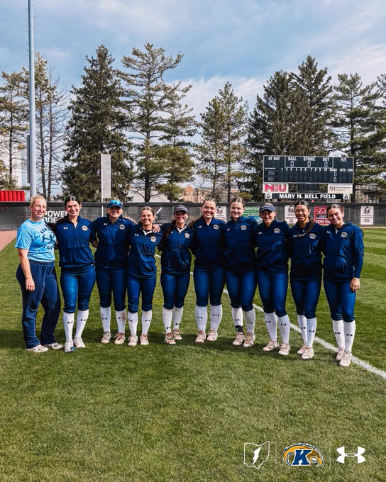 Eleven Kent State softball players and a coach pose in a line on the field at Mary M. Bell Field, smiling for a group photo. The NIU scoreboard showing KSU vs. NIU is visible in the background.