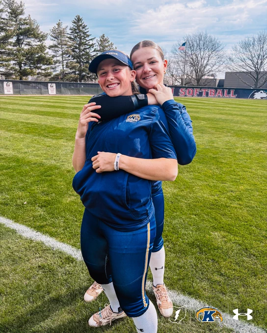 Two Kent State softball players in navy uniforms embrace and smile on the field at NIU, with the 'NIU Softball' outfield wall visible in the background.