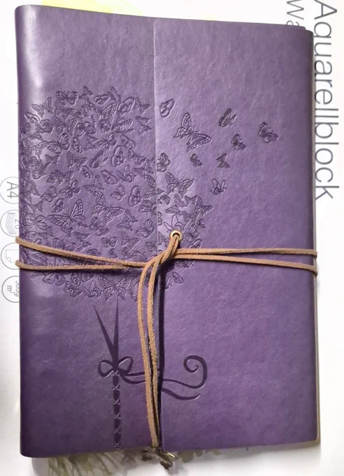 Mein lila Buch, in das ich meine Gedichte schreibe, liegt auf einem Aquarell Block.
Das Buch wird mit einem Band verschlossen, an dem am Ende ein Schmetterling hängt. Auf dem Buch sind viele Schmetterlinge. 