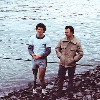 Terry Fox stands on a rocky shoreline beside a body of water, wearing a light blue athletic shirt with darker sleeves, short running shorts, white socks, and running shoes. His right leg is a prosthetic. Doug Alward stands next to him in a tan jacket and dark pants, hands in his pockets. A rope or cable runs along the edge of the shore in front of them. The water extends into the background with a distant shoreline and buildings visible across it.
