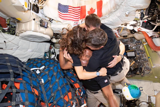 Artemis II crew members share a joyful hug aboard the Orion spacecraft, with American and Canadian flags visible in the background. 