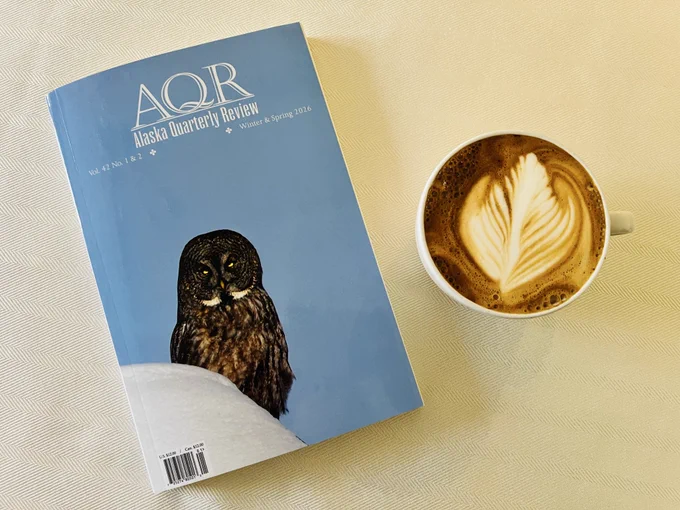 Pale blue cover with owl—Alaska Quarterly Review. Beside a cappuccino.