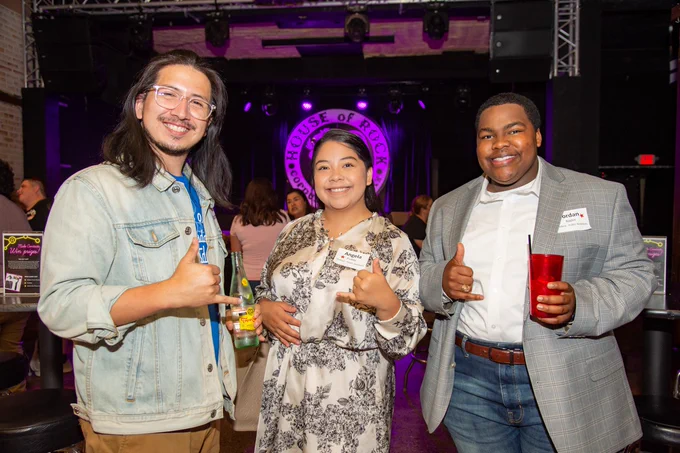 A group of TAMU-CC young alumni smiling and making a shakas up hand gesture while standing in front of a "House of Rock" sign inside a venue. They are holding drinks and wearing name tags.