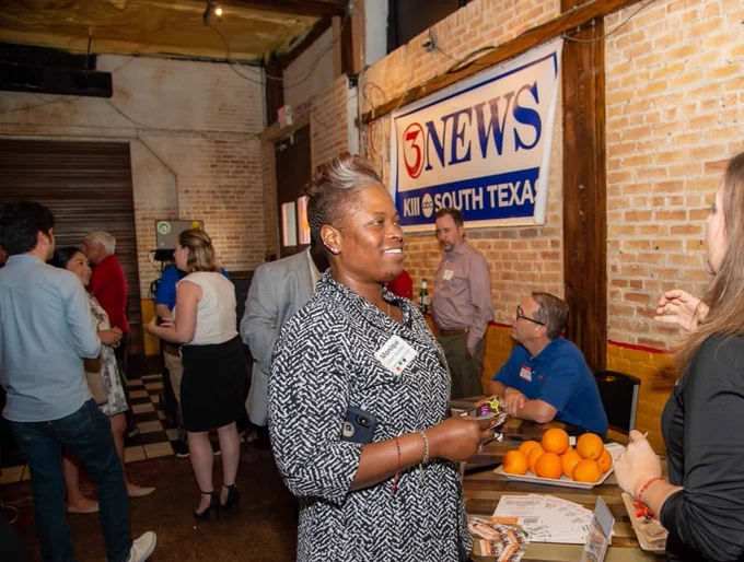 A group of people are socializing at an indoor TAMU-CC alumni professionals networking event. A sign for "3 NEWS KIII SOUTH TEXAS" is displayed on the wall. A table has oranges and flyers near seated individuals engaged in conversation. The setting includes exposed brick walls and a checkerboard floor.