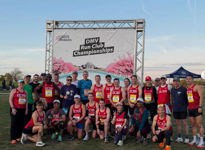the 2026 DC Road Runners Cherry Blossom DMV Run Club Team posing in front of a Cherry Blossom Ten Miler banner in the early morning sunrise