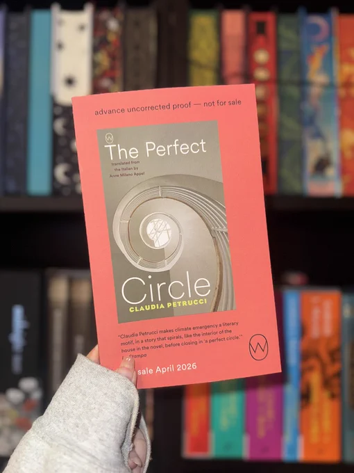 A person holds an orange book titled "The Perfect Circle" in front of a colorful bookshelf. The book is an advance proof.