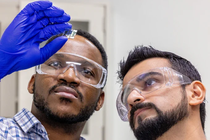 Two men with dark short hair and facial hair wear protective eyewear and gloves. One of them is holding a small rectangular solar cell.