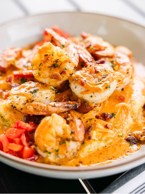 A dish featuring shrimp placed on a bed of creamy grits, garnished with chopped red peppers and herbs. The shrimp are coated in a rich, orange sauce.