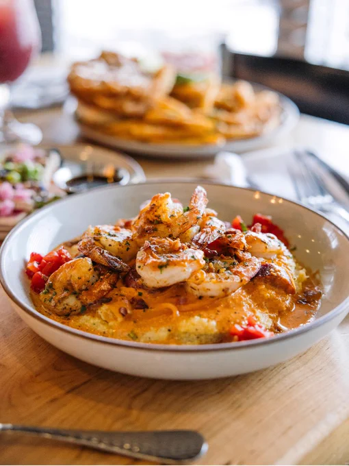 A bowl of shrimp and grits garnished with herbs and red peppers is placed on a wooden table. In the background, there is another dish, likely nachos, with a blurred effect. A drink is partially visible on the left.