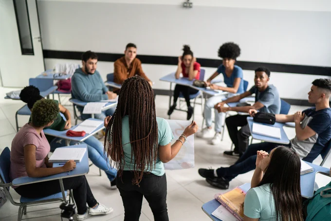 A group of students is seated in a semicircle in a classroom, attentively listening to a person standing and speaking. Desks are arranged in a U-shape. The room is brightly lit and the atmosphere appears engaged and focused.