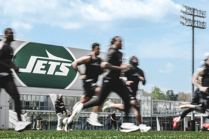 Athletes running on a green field in front of a New York Jets facility, with blurred faces and a large Jets logo visible.