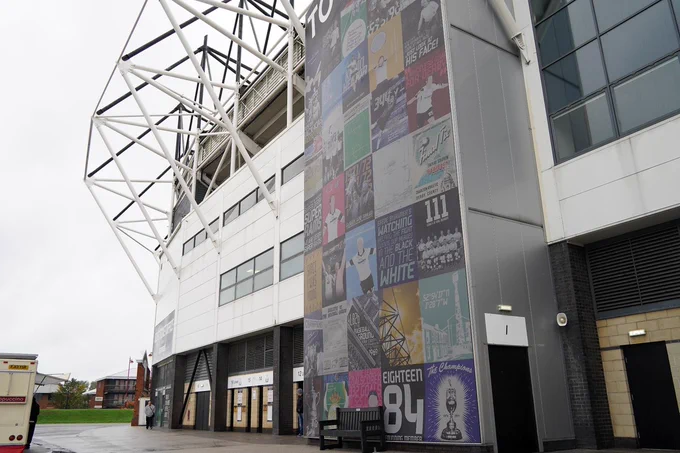 The image shows an exterior wall adorned with a colorful mural composed of various square tiles, each displaying different Derby County designs and texts. The stadium's architecture and a cloudy sky are in the background.