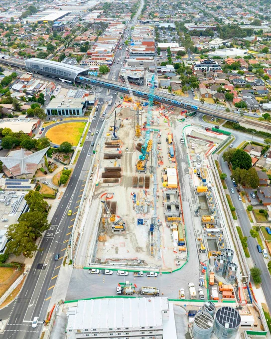 Aerial view of a Suburban Rail Loop site in Clayton, with a railway station and tracks running through it. The site is busy with various construction vehicles and equipment. Buildings, roads, and residential houses surround the construction site.