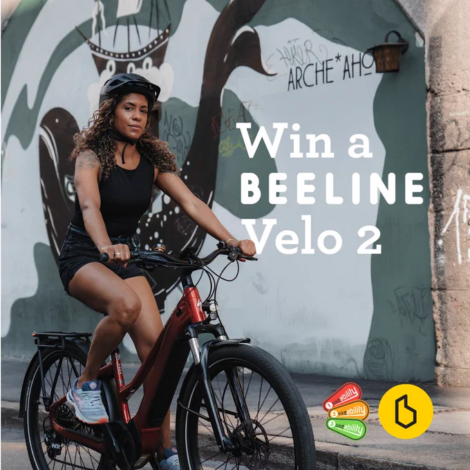 Woman riding a red electric bike against a graffiti-covered wall with 'Win a BEELINE Velo 2' text overlay.