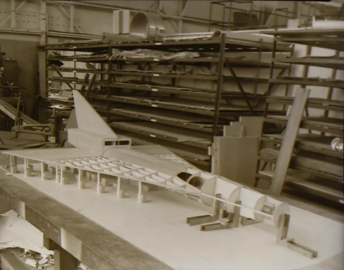A detailed scale model of a jet aircraft under construction in a workshop filled with shelves and materials.