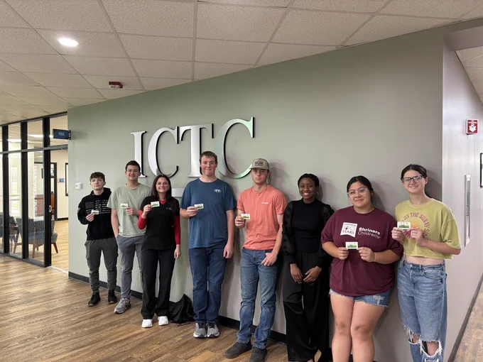 A group of individuals stands in a room with a light green wall featuring the letters ICTC.