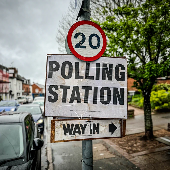 A weathered street sign reading “POLLING STATION” with a smaller “WAY IN” arrow pointing right, mounted below a 20 mph speed limit sign on a rainy residential street with parked cars and trees in the background.