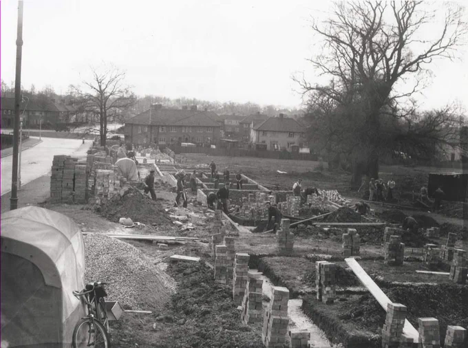 A black and white photo of a building site. A bicycle leans against a shed to the left, and piles of bricks are stacked next to foundations. There are a couple of trees and other houses in the background.