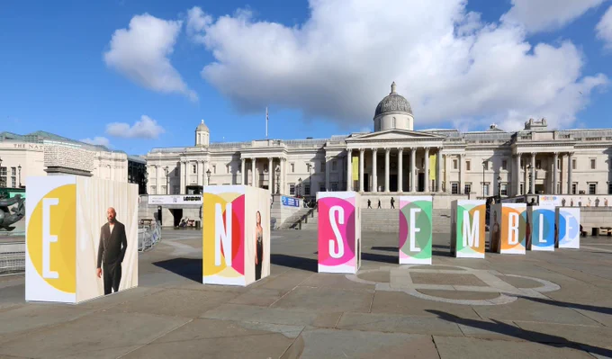 A row of large freestanding colourful panels spelling out “ENSEMBLE” is arranged across Trafalgar Square, with each panel also featuring a portrait of a featured participant. The National Gallery forms the backdrop under a blue sky with scattered clouds.