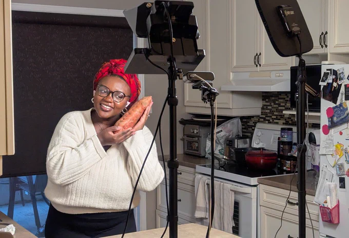 Pink cradling a sweet potato in a kitchen. She is facing a webcam, a prompter and a key light.