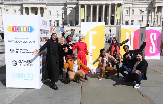 Deputy Mayor Justine Simons OBE, stands with a group of people posinh together in Trafalgar Square in front of large colourful “ENSEMBLE” installations at the London Games Festival launch. A tall display panel beside them shows the Ensemble branding and logos for the Mayor of London, UK Government funding, Games London and Film London.