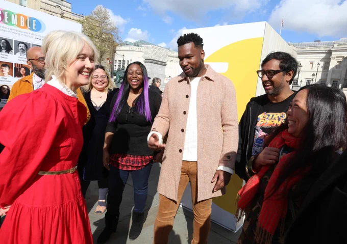 Deputy Mayor Justine Simons OBE speaks with a small group of people at the London Games Festival launch in Trafalgar Square. The group stands in front of the large yellow “E” panel from the ENSEMBLE installation, with festival signage visible behind them.