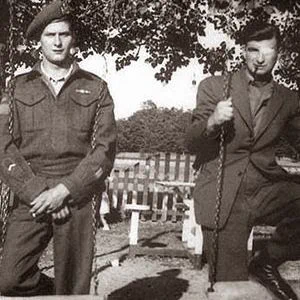 Wilfrid Arsenault and Léo Major stand on a wooden swing set beneath a leafy tree. Wilfrid Arsenault, on the left, wears a military uniform with a beret, standing with one foot on the swing’s lower bar and his hands clasped in front. Léo Major, on the right, wears a suit and tie, holding the swing rope with one hand while resting a foot on the lower bar. A wooden fence and open yard are visible behind them, with sunlight creating clear shadows on the ground.
